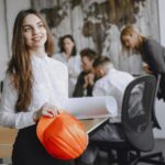 Smiling businesswoman with a hard hat in a modern office setting, engaging with colleagues.